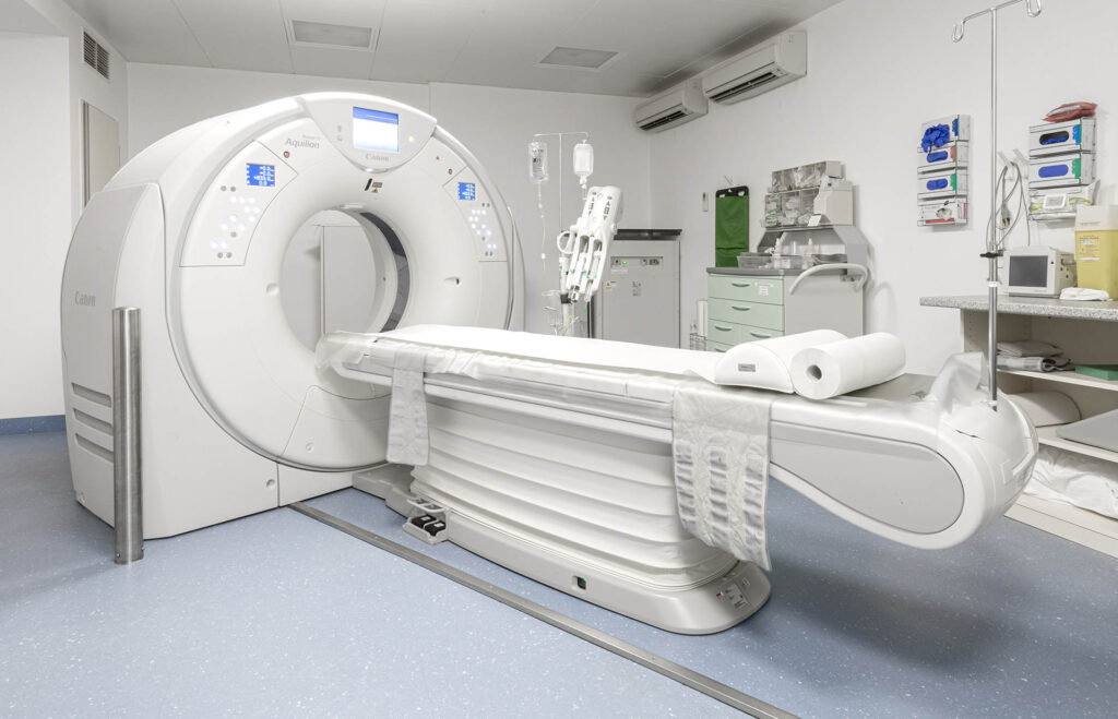 Ein CT-Scanner in einem sauberen, gut beleuchteten medizinischen Raum, in dem im Hintergrund medizinische Geräte, Versorgungsschubladen und Monitore zu sehen sind.