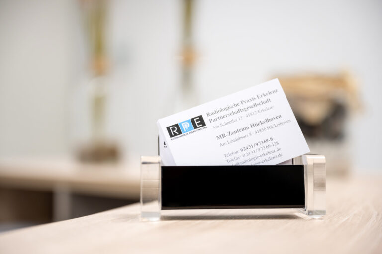 Ein Visitenkartenhalter mit einer sichtbaren Visitenkarte der radiologischen Praxis Erkelenz liegt auf einer gesprenkelten Arbeitsplatte. Dekorative, blumenförmige Objekte sind im Hintergrund unscharf.