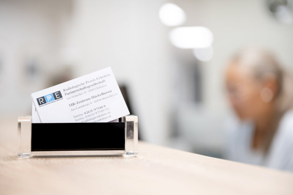 Ein Visitenkartenhalter mit einer sichtbaren Visitenkarte der radiologischen Praxis Erkelenz liegt auf einer gesprenkelten Arbeitsplatte.