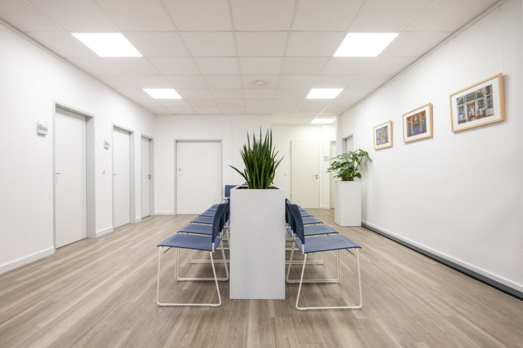 Modernes Wartezimmer mit blauen Stuhlreihen, einem zentralen Pflanzgefäß mit einer hohen Pflanze, gerahmten Bildern an der Wand und mehreren geschlossenen Türen.