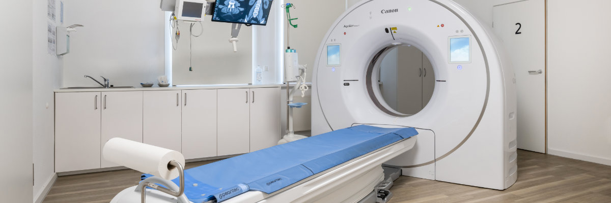Ein modernes CT-Gerät mit einem blauen Patiententisch steht in einem sauberen, hellen medizinischen Untersuchungsraum mit Monitoren und Schränken.